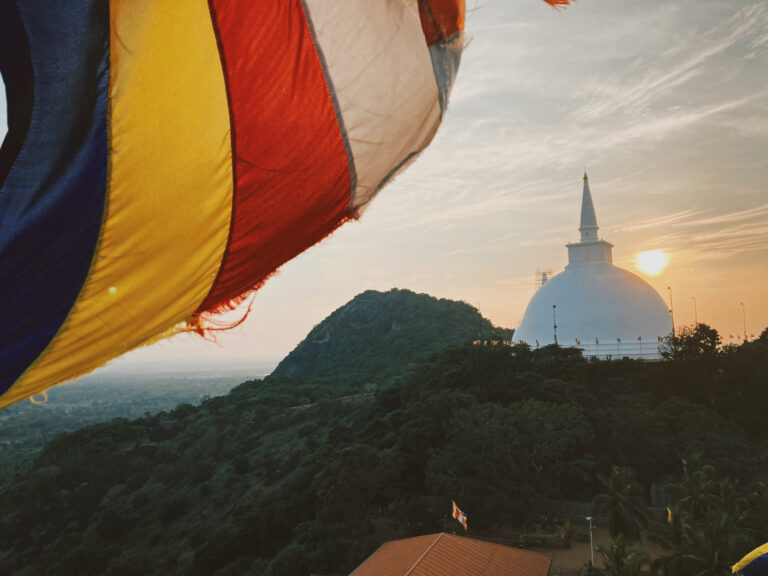 A white stupa on top of a mountain covered by trees, at sunset.a third of the image is taken by a buddhist flag, waving very close to the camera.