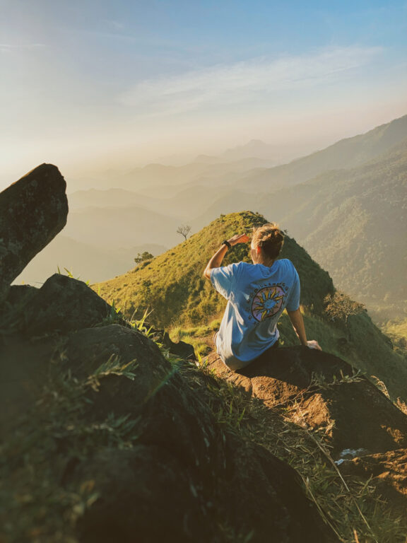 A young female sitting on a rock on top of a mountain, at sunrise, looking towards the sun, with her hand raised over her eyes. There are many layers of mountains covered in vegetation in the background.