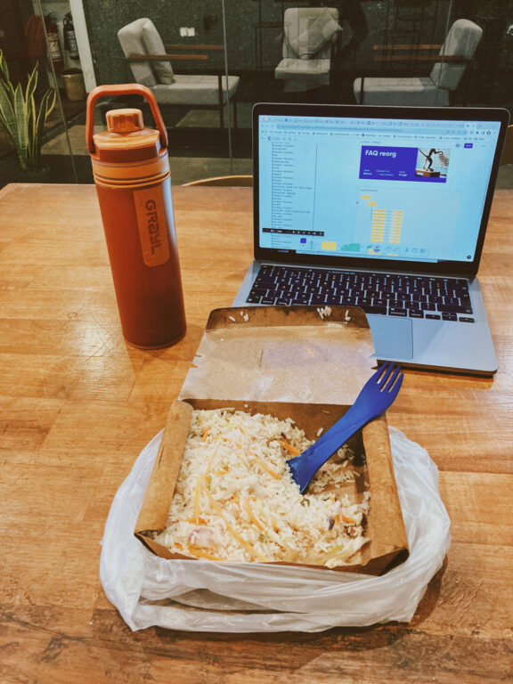 A water bottle, an open laptop with a work tool open on desktop and a takeaway box of rice on a desk in a hostel lounge.