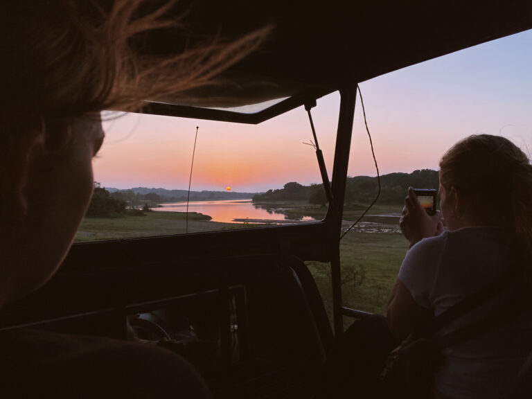 Natural landscape featuring a lake at sunrise. The photo is taken from inside a jeep and we can see its metal bars and roof, framing the landscape.On the right a tourist sat on the jeep is taking a photo of the landscape. On the left we see part of the face of another tourist, looking at the sunrise.