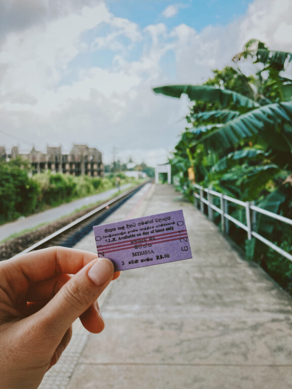 In the foreground:A hand holding a third class train ticketIn the background, blurred: a train platform. On the left we can see the railway and on the right there are some banana trees. The sky is cloudy.