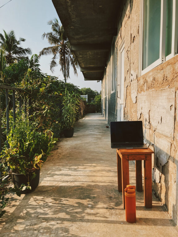 A balcony with a small stool on which sits an open laptop. The house walls on the right are ruined and the plants in the garden on the left are thriving. There's a warm sunset light.