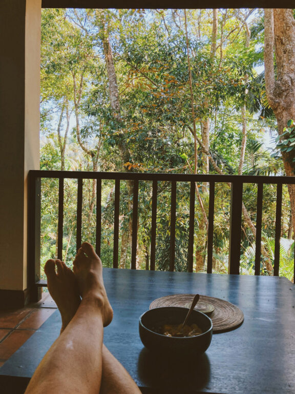 A short table on a balcony, overlooking some tall trees on a sunny day.There's a breakfast bowl on the table, with a spoon, and the person who took the photo has her feet on the table, with her legs crossed.