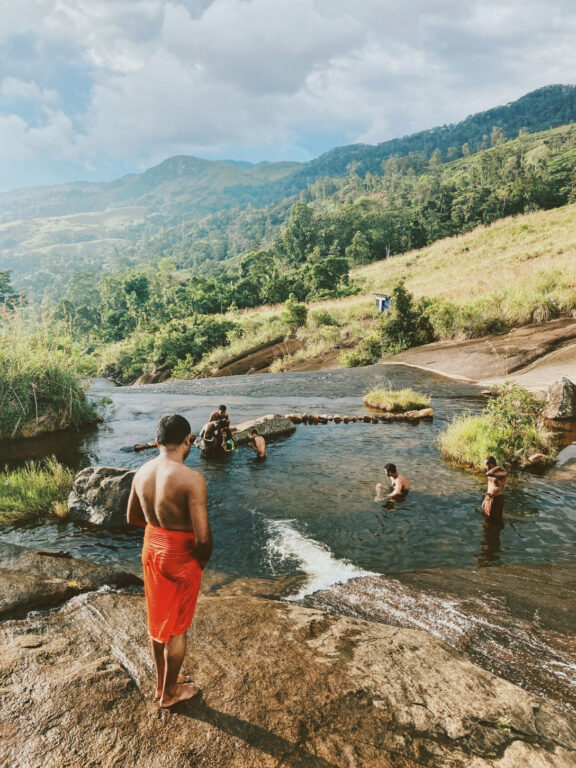 A natural setting in what seems a remote location, with a small waterfall and some green hills in the background, at sunset.A monk in an orange dress is standing in the foreground, looking down the rock where he's standing, at the other people who are bathing at the bottom, in a natural pool.