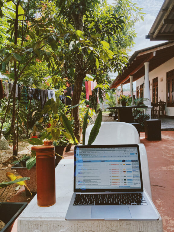 A water bottle and a laptop on a small table in front of a modest house. The laptop shows a spreadsheet. The House is surrounded by a garden with plants and some laundry that has been hung to dry.