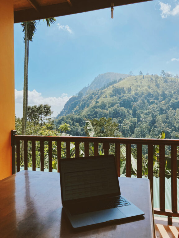 A table on a balcony that overlooks a hill covered in trees on a sunny day. The balcony is in the shade and there's an open laptop on it.
