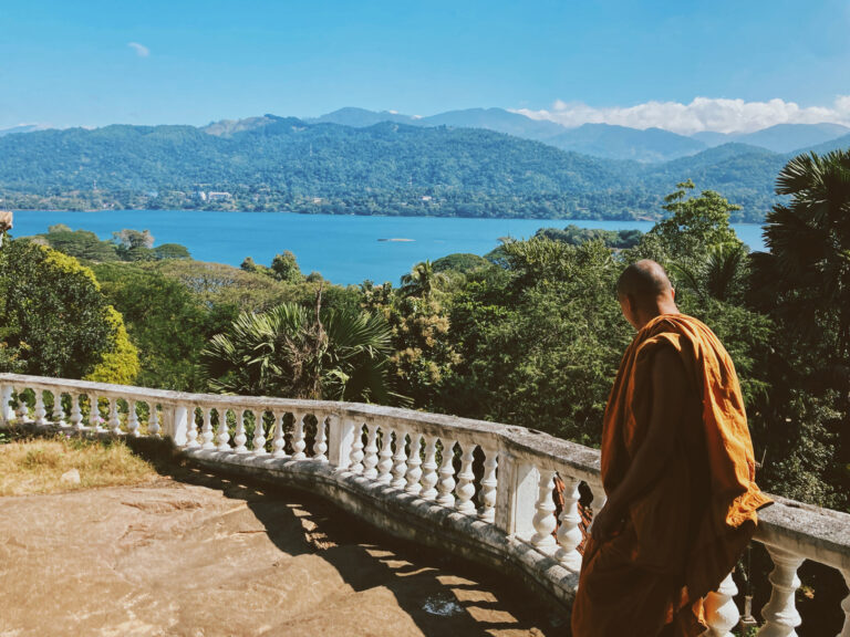 A monk dressed in orange, sat on a cement banister of a large terrace looking at the view made of a blue lake and some green hills on a sunny day.